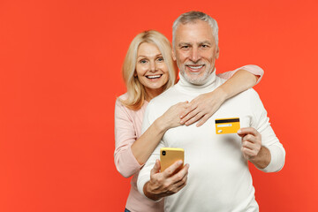 Cheerful couple two friends elderly gray-haired man blonde woman in white pink clothes using mobile cell phone hold credit bank card hugging isolated on bright orange color background studio portrait.