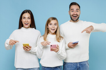 Wall Mural - Excited young parents mom dad with child kid daughter teen girl in white sweaters point on mobile phone typing sms message isolated on blue background studio. Family day parenthood childhood concept.