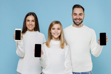 Wall Mural - Smiling young parents mom dad with child kid daughter teen girl in white sweaters hold mobile phone with blank empty screen isolated on blue background studio. Family day parenthood childhood concept.