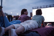 Two girls lying on the roof of a car, having fun while watching a movie in an open air cinema with a big white screen