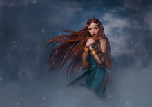 Redhead Goddess Fantasy Woman Walks In The Clouds. Fashion Model Posing In Studio Background Dramatic Winter Sky With Smoke. Elf Princess Girl. Long Red Hair Flying In Wind Snow Is Falling. Blue Dress
