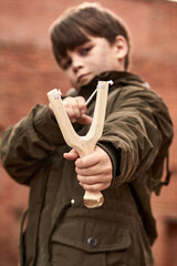 child boy holding slingshot in hands, going to play, shoot. point at camera, in city street. focus on wooden slingshot