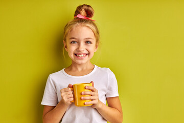 cheerful kid girl drink tea before school, she is joyful, looks at camera smiling isolated over green background