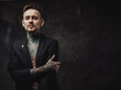 © Fxquadro - Dressed in black luxurious jacket serious and tattooed hipster person with modern haircut and short beard poses in dark background.