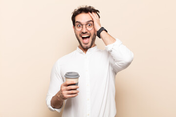 young handsome man raising hands to head, open-mouthed, feeling extremely lucky, surprised, excited and happy