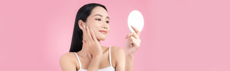 Wall Mural - Attractive young Asian woman holding mirror and feeling happy about her skin. Isolated on pink background