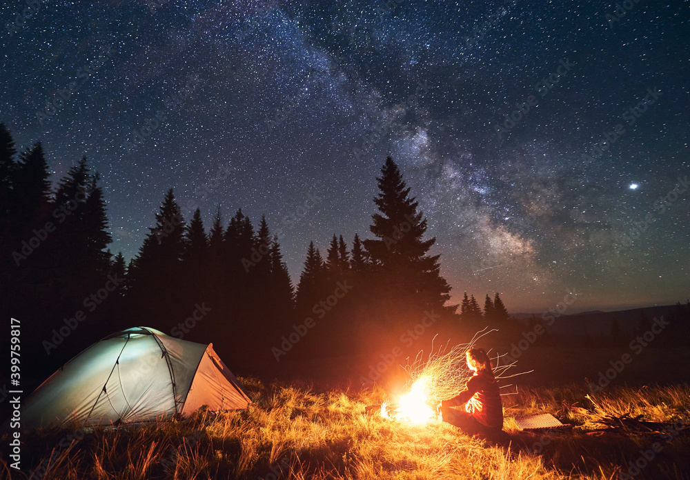 Girl is sitting by the fire on background of tent and spruce forest under starry sky on which milky way is visible. Bonfire with a big flame under the night sky - obrazy, fototapety, plakaty 