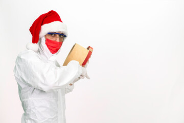 Wall Mural - Health worker wearing protective clothing. He wears a santa hat on his head. He has a red corona mask on his face. He is holding a gift box in the shape of a heart.