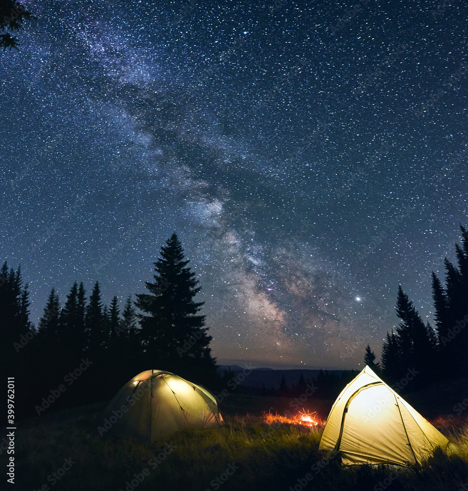 Night camp with burning bonfire and two tents in pine forest under bright starry sky on which Milky Way is clearly visible. In distance you can see valley of hills and mountains. Mighty landscape - obrazy, fototapety, plakaty 