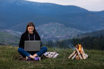 young female freelancer working on laptop in the mountains in the evening. Tourist girl sitting near campfire and having fun. Copy space.