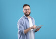 © Prostock-studio - Positive young guy in casual wear using tablet computer over blue studio background