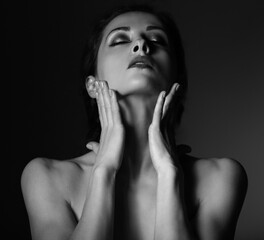 Beautiful mysterious woman in darkness with healthy neck, shoulders and touching her face skin on dramatic  black background with empty copy space. Closeup portrait