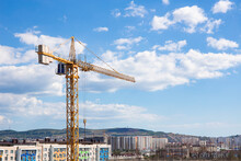 Yellow Construction Crane Or Tower With A Cabin At Height, Against The Backdrop Of A Clear Blue Sky And Cityscape