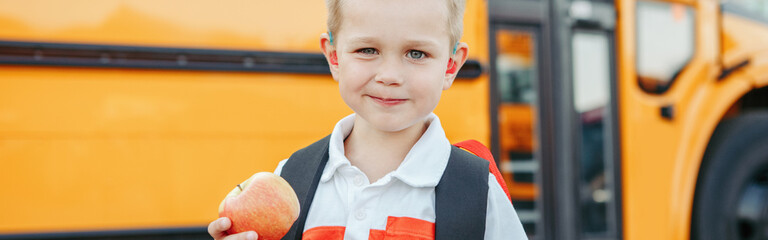 Caucasian boy student with backpack near yellow bus on first September day. Child kid eating apple at school yard outdoors. Education and back to school in Autumn Fall. Web banner header.