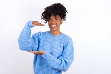 Wall Mural - oung beautiful African American woman wearing blue knitted sweater against white wall gesturing with hands showing big and large size sign, measure symbol. Smiling looking at the camera.