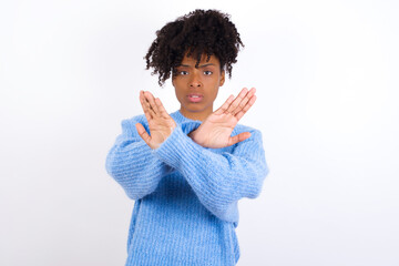 Wall Mural - Young beautiful African American woman wearing blue knitted sweater against white wall has rejection expression crossing arms and palms doing negative sign, angry face.