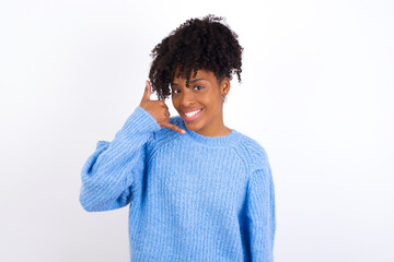 Wall Mural - Young beautiful African American woman wearing blue knitted sweater against white wall smiling doing phone gesture with hand and fingers like talking on the telephone. Communicating concepts.