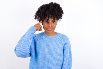 Wall Mural - Young beautiful African American woman wearing blue knitted sweater against white wall tries to memorize something, keeps fore finger on temple, reminds information for exam,