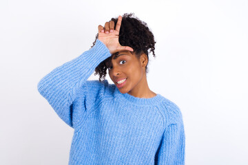 Wall Mural - Young beautiful African American woman wearing blue knitted sweater against white wall making fun of people with fingers on forehead doing loser gesture mocking and insulting.