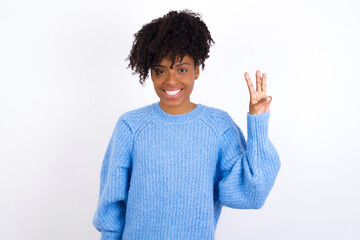 Wall Mural - Young beautiful African American woman wearing blue knitted sweater against white wall showing and pointing up with fingers number three while smiling confident and happy.