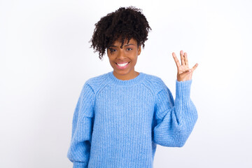 Wall Mural - Young beautiful African American woman wearing blue knitted sweater against white wall showing and pointing up with fingers number four while smiling confident and happy.