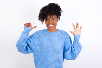 Wall Mural - Young beautiful African American woman wearing blue knitted sweater against white wall showing and pointing up with fingers number six while smiling confident and happy.