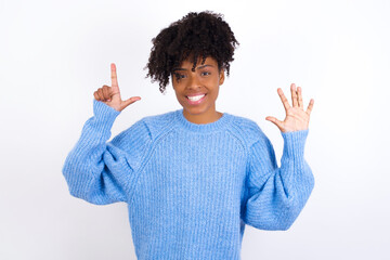 Wall Mural - Young beautiful African American woman wearing blue knitted sweater against white wall showing and pointing up with fingers number seven while smiling confident and happy.