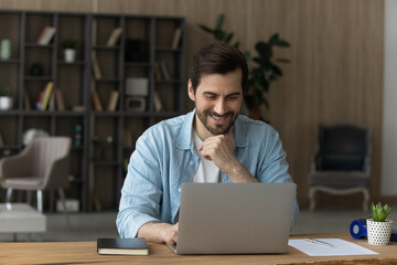 Smiling millennial male sit at home office desk look at laptop screen work distant on gadget. Happy young man use computer, browse wireless internet on device. Online communication concept.