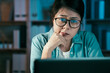 © PR Image Factory - Worrired young asian korean female freelancer with eyeglasses holding hand on chin while sitting in home office late at night in front of laptop computer. frowning girl worker staring at screen.