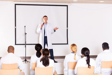 Canvas Print - Confident male speaker in white coat giving presentation from stage at medical conference