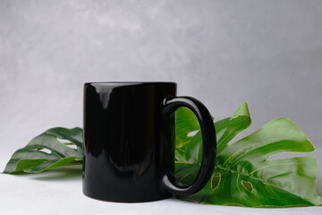 Black mug mock up. Ceramic mug and plant leaves on grey background. Blank coffee or tea mug.