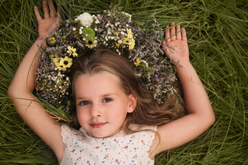 Wall Mural - Cute little girl wearing wreath made of beautiful flowers on green grass, top view