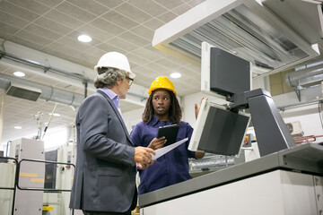 Wall Mural - Black female factory worker and her male boss standing at industrial machine and talking. Low angle. Production process or machinery concept