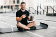 © Ivan - Man resting after outdoors workout with water and phone. Healthcare concept. Young sporty man after practicing yoga, break in doing exercise, relaxing on yoga mat, texting on smartphone.