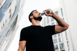 © Ivan - Athletic man drinking water from plastic bottle after jogging. Sporty male runner with dark short hair. Black t-shirt. Blurred apartment blocks on background. Low angle shot. Drink more water.