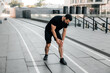 © Ivan - Attractive man feeling pain in his foot during sport workout outdoors. Male runner in black sportswear and sneakers standing and feeling cramps in his left leg. Urban sport concept.
