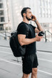 © Ivan - Friendly man in black clothes talking on the phone, smiling and looking away. Man standing near apartment blocks, holding black sports bottle. Urban life concept.