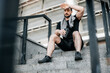 © Ivan - Resting time A man in black clothes with towel on his shoulders sits at the top of the stairs and rests after a hard jog or workout and wipes sweat with his hand. Morning fitness. City on background.