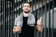 © Ivan - Cheerful man with smartphone in hand thumbing up. Drink more water. Urban sport concept. Man with towel on his shoulders resting after hard morning workout. Jogging concept.