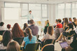 © kasto - Business and entrepreneurship symposium. Speaker giving a talk at business meeting. Audience in conference hall. Rear view of unrecognized participant in audience.