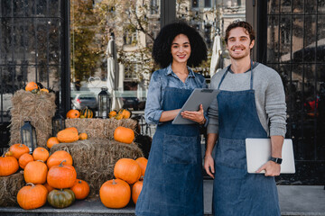 Happy young coffee shop owners standing outdoor near their cafe with laptop and decorating pumpkins preparing for halloween on background.