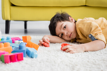 Boy lying on floor near colorful building blocks on carpet on blurred foreground