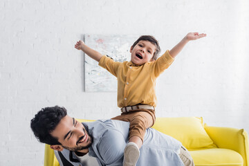 Wall Mural - Cheerful boy sitting on back of hispanic father in living room, two generations of men