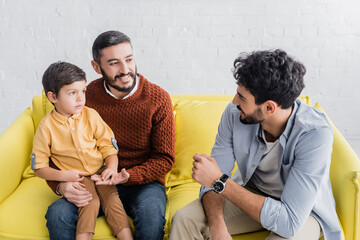 Wall Mural - Smiling hispanic man hugging grandson near son on couch at home, three generations of men