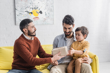 Wall Mural - Hispanic man giving present to grandson near smiling father at home, three generations of men