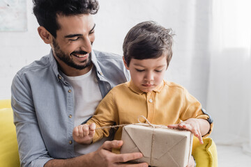 Wall Mural - Boy unpacking gift box near cheerful hispanic father, two generations of men