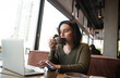 © Ivan - Freelancer working in cozy atmosphere concept. Concentrated young woman drinks coffee from grey cup and looks into laptop on her table with attention. Big well illuminated windows on background.