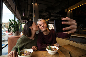 Man and woman in love take photo together in a public place. Attractive brunette woman kisses her boyfriend's cheek while he holds smartphone to take selfie. Social media concept. Cafe of restaurant.