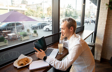Young freelancer happy with a phone call. Remote work concept. Coffee house on background. Tasty croissant and cup of latte. Male freelancer programmer working in a coffee shop.