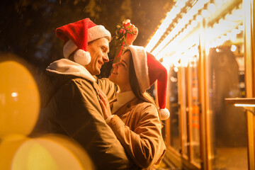 Wall Mural - Happy couple in Santa hats standing under mistletoe bunch outdoors, bokeh effect